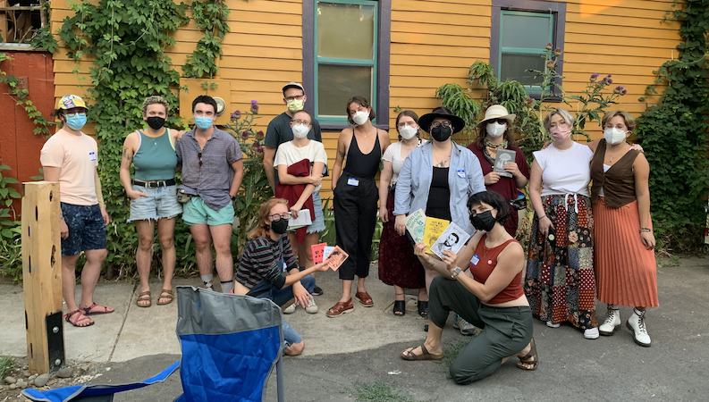 A group of 13 people wearing masks, several of whom are holding zines. They're standing in front of a yellow wall with two windows framed in blue. A blue chair is in the foreground.