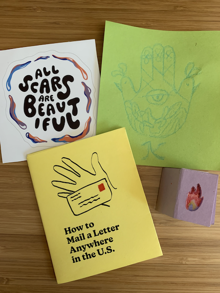 A sticker reading "All Scars are Beautiful", a hamsa drawn on green paper, a yellow zine titled "How to mail a letter anywhere in the US", and a tiny purple zine with a flame on the cover